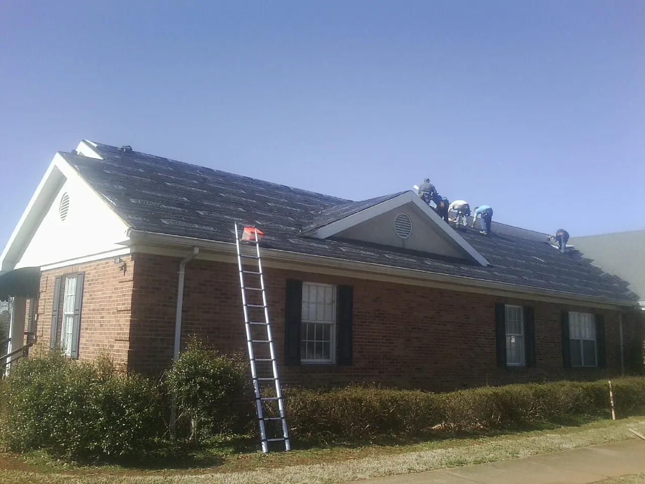 Crew tearing shingles off a brick ranch roof with a ladder set — Emergency Roof Repair work by our crew in Clanton