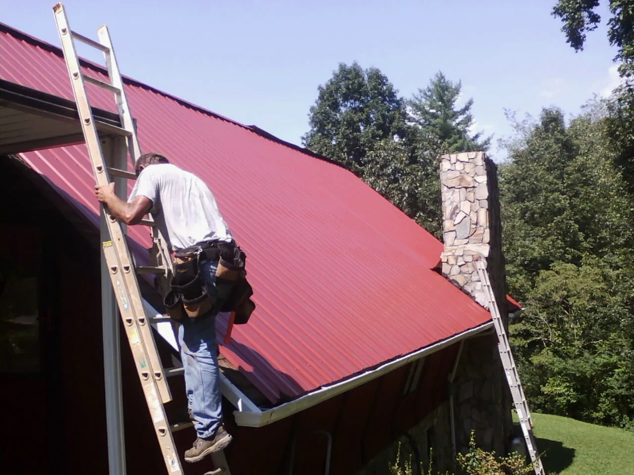 Roofer on a ladder beside a red metal roof and stone chimney — Soffit & Fascia Repair by Roofing Brothers in Clanton